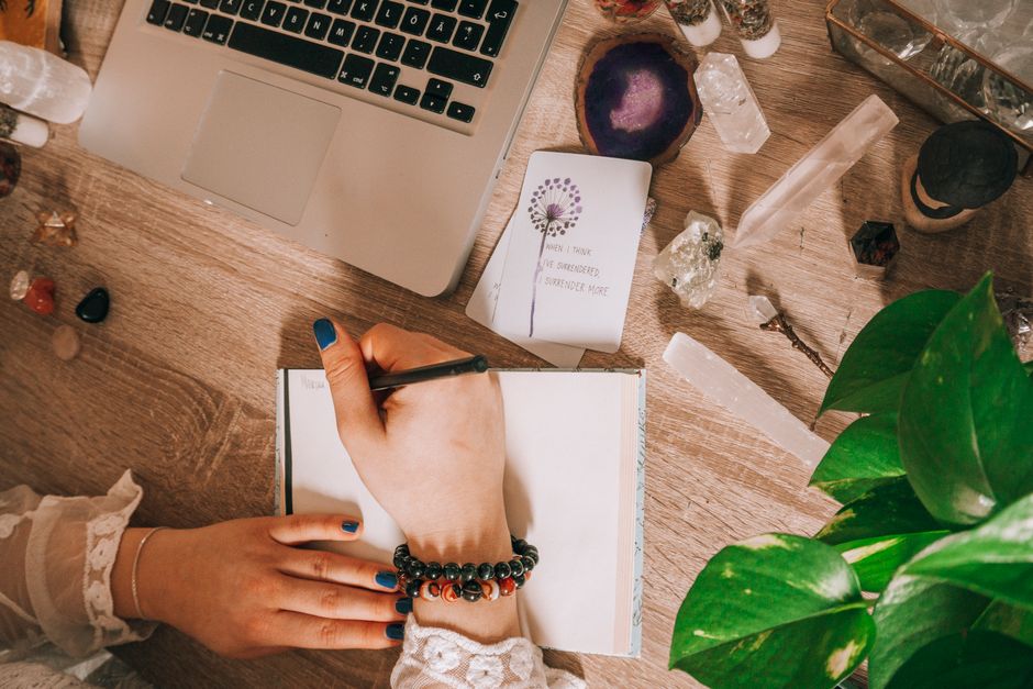 mental wellness news - Woman's hand writing in a journal surrounded by crystals, plants, and laptop on a wooden desk.