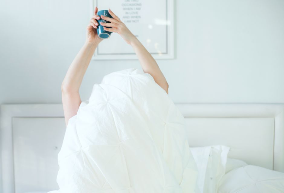 mental wellness - A person holding a coffee mug while relaxing in bed, covered with a white sheet.